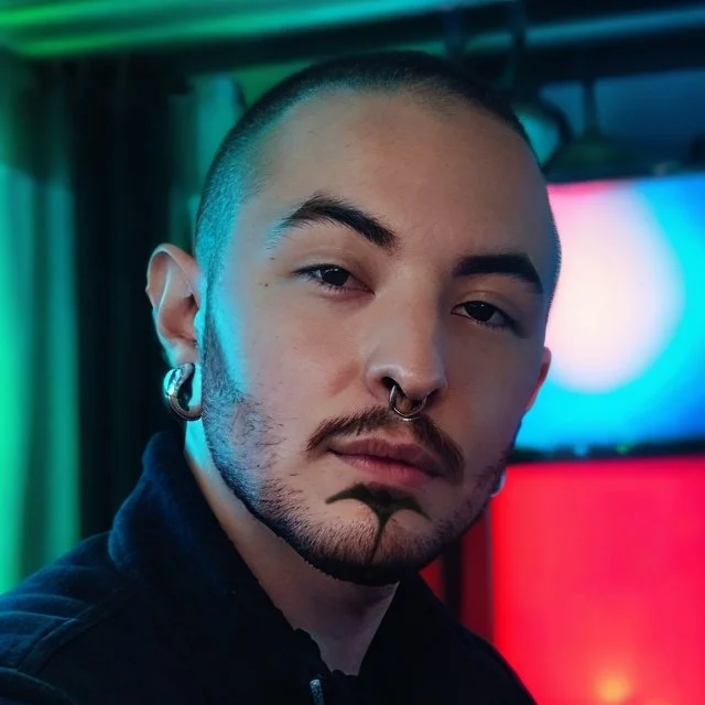 A close-up headshot of Kato Lujan Camacho, a man with a shaved head, a trimmed beard with a unique chin design, a septum piercing, and stretched earlobes, set against a colorful, softly blurred background.